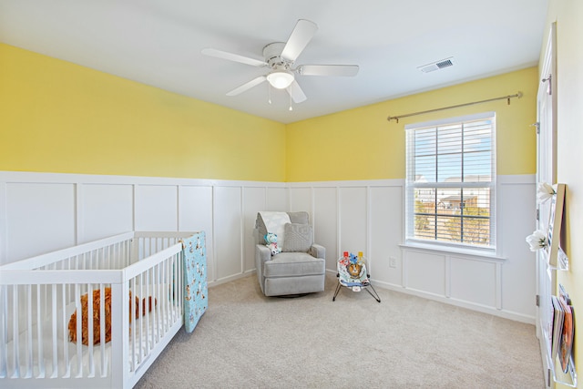 bedroom with a crib, visible vents, wainscoting, ceiling fan, and carpet floors