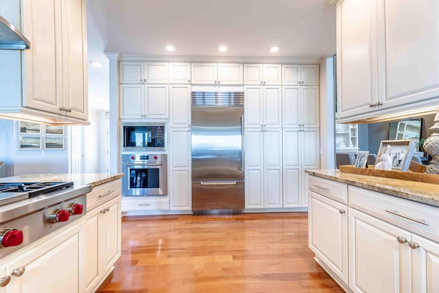kitchen featuring white cabinets, built in appliances, light wood-style flooring, and wall chimney exhaust hood