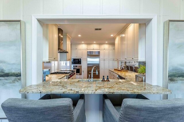 kitchen with a sink, wall chimney exhaust hood, built in appliances, and light stone counters