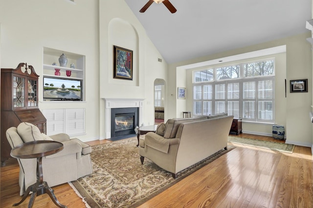 living room featuring arched walkways, high vaulted ceiling, a fireplace with flush hearth, wood finished floors, and baseboards