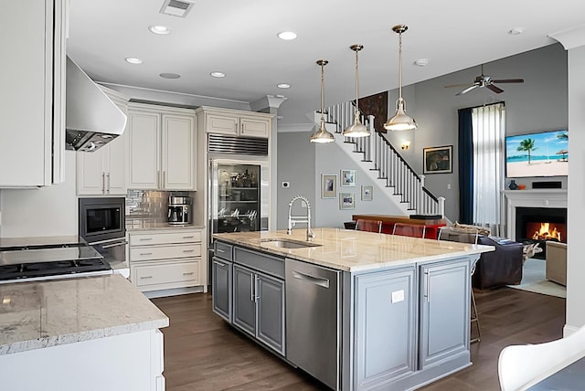 kitchen featuring sink, wall chimney exhaust hood, hanging light fixtures, built in appliances, and a kitchen island with sink