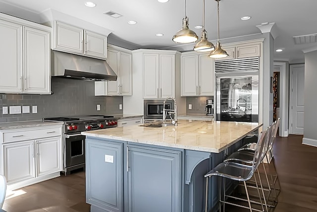 kitchen with light stone countertops, sink, built in appliances, hanging light fixtures, and an island with sink