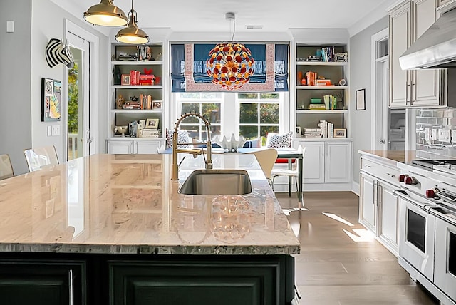 kitchen featuring gas stove, sink, a center island with sink, hanging light fixtures, and range hood