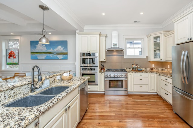 kitchen with light stone counters, light wood-style floors, stainless steel appliances, wall chimney exhaust hood, and a sink