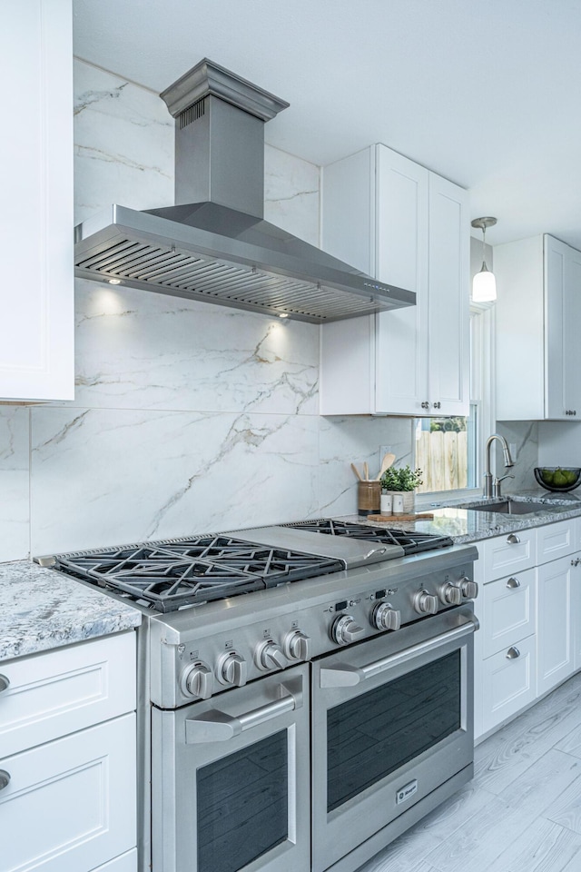 kitchen with wall chimney range hood, double oven range, light stone counters, white cabinetry, and a sink