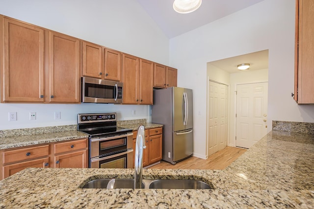 kitchen featuring brown cabinets, a sink, light stone counters, stainless steel appliances, and vaulted ceiling