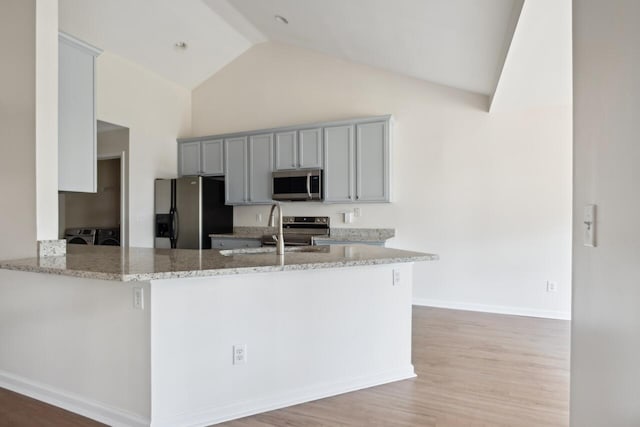 kitchen with stainless steel appliances, a sink, gray cabinetry, light stone counters, and high vaulted ceiling
