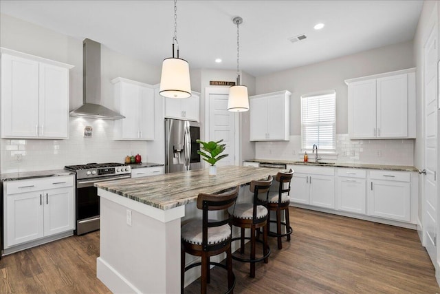 kitchen with appliances with stainless steel finishes, decorative light fixtures, white cabinets, a center island, and wall chimney range hood