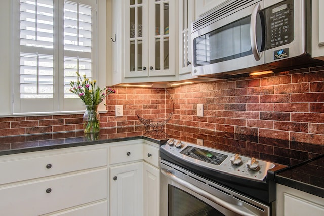 kitchen featuring white cabinets, dark countertops, glass insert cabinets, stainless steel appliances, and backsplash