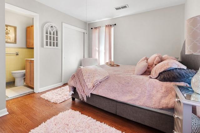 bedroom featuring visible vents, a wainscoted wall, wood finished floors, ensuite bathroom, and tile walls