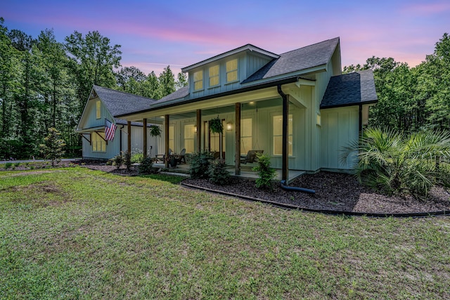 back of property at dusk featuring board and batten siding, a lawn, a shingled roof, and covered porch
