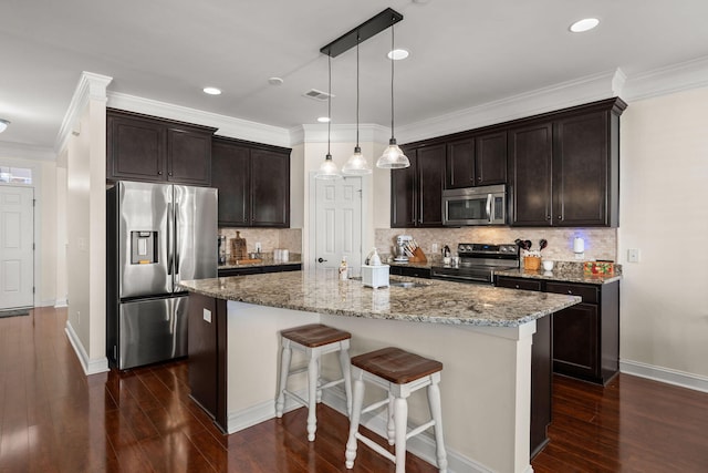 kitchen featuring backsplash, stainless steel appliances, light stone counters, an island with sink, and decorative light fixtures