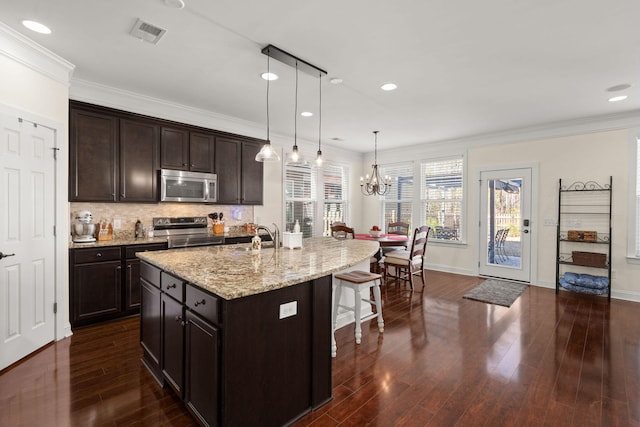 kitchen with crown molding, appliances with stainless steel finishes, dark brown cabinetry, an island with sink, and decorative light fixtures