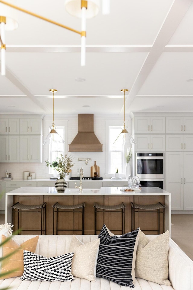 kitchen with light countertops, custom range hood, decorative light fixtures, and a kitchen island with sink