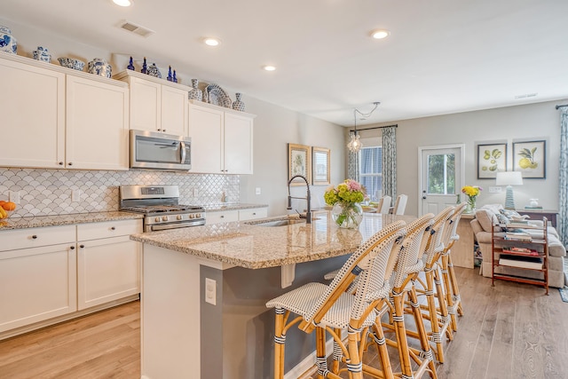 kitchen featuring stainless steel appliances, a sink, hanging light fixtures, light stone countertops, and a center island with sink