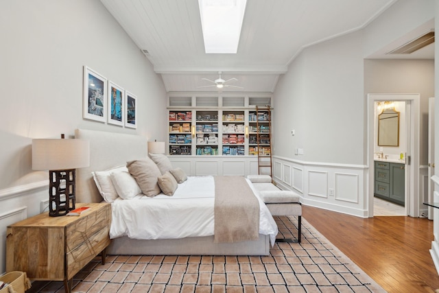 bedroom with vaulted ceiling with skylight, a wainscoted wall, ensuite bath, wood finished floors, and a decorative wall