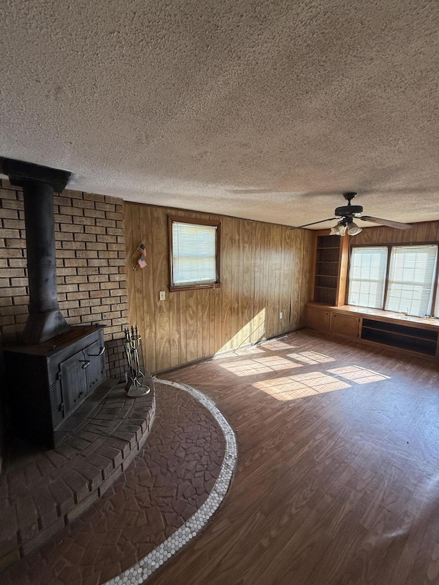 unfurnished living room with a wood stove, a textured ceiling, wood walls, a ceiling fan, and wood finished floors