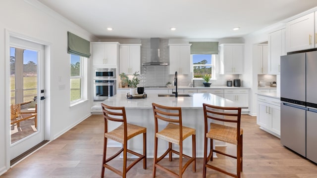 kitchen with wall chimney exhaust hood, sink, white cabinetry, an island with sink, and stainless steel appliances