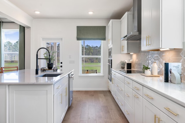 kitchen with wall chimney range hood, appliances with stainless steel finishes, a kitchen island with sink, white cabinetry, and backsplash