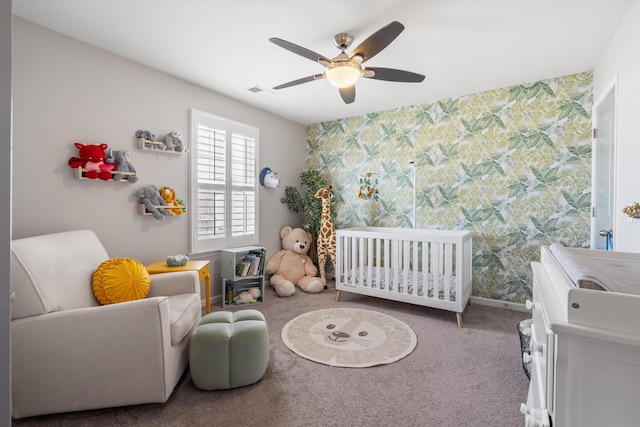 carpeted bedroom with a crib, wallpapered walls, baseboards, visible vents, and a ceiling fan