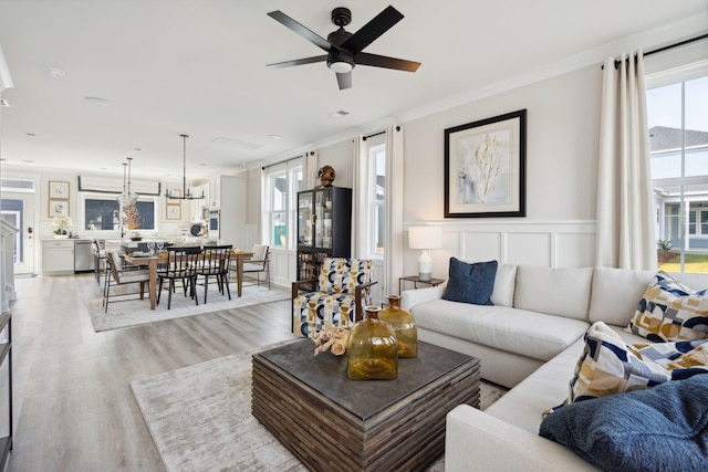 living room featuring light wood-style flooring, a decorative wall, a wainscoted wall, a ceiling fan, and ornamental molding