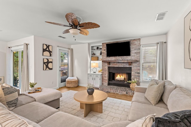 living room with a brick fireplace, light hardwood / wood-style flooring, ceiling fan, and built in shelves