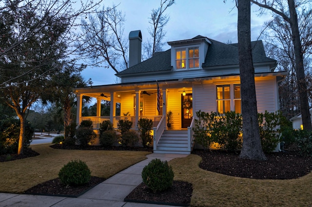 view of front facade featuring covered porch, a shingled roof, a ceiling fan, a front lawn, and a chimney