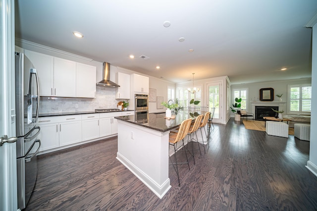 kitchen featuring a kitchen island with sink, wall chimney exhaust hood, stainless steel appliances, and white cabinets