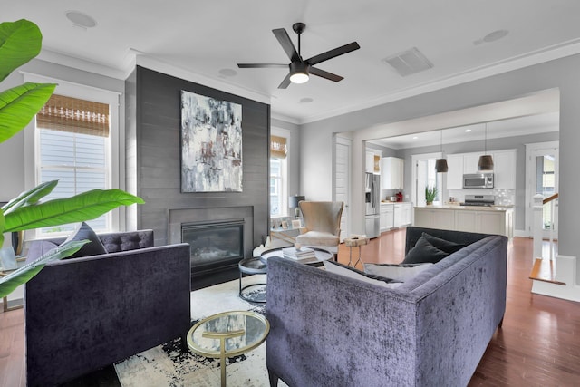 living room with visible vents, ornamental molding, a glass covered fireplace, ceiling fan, and wood finished floors