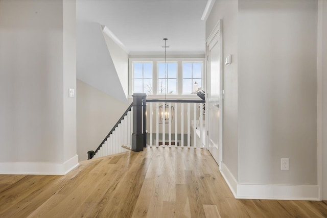 hallway with a notable chandelier, light wood-style floors, ornamental molding, an upstairs landing, and baseboards