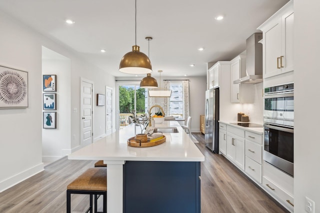 kitchen featuring a sink, wall chimney range hood, stainless steel appliances, light countertops, and a kitchen island with sink