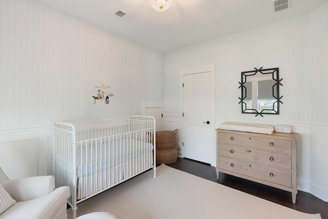 bedroom with a wainscoted wall, a nursery area, dark wood-style floors, and visible vents