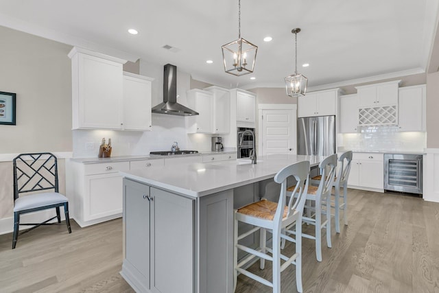 kitchen with appliances with stainless steel finishes, an island with sink, white cabinets, wine cooler, and wall chimney exhaust hood