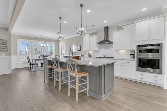 kitchen featuring appliances with stainless steel finishes, white cabinets, hanging light fixtures, a center island with sink, and wall chimney exhaust hood