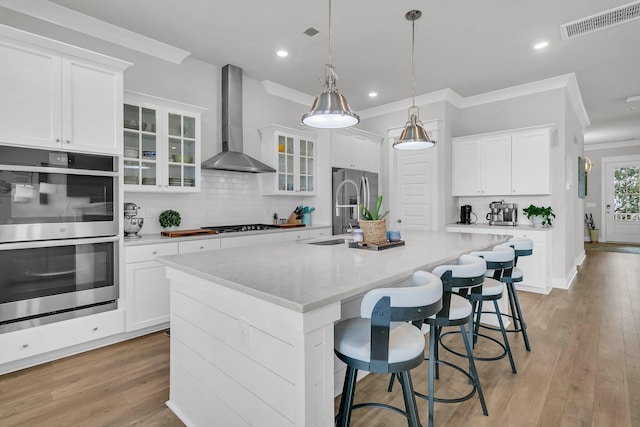 kitchen with white cabinetry, a kitchen island with sink, stainless steel appliances, wall chimney range hood, and decorative backsplash