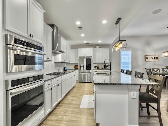 kitchen featuring sink, a kitchen island with sink, wall chimney range hood, stainless steel appliances, and a breakfast bar