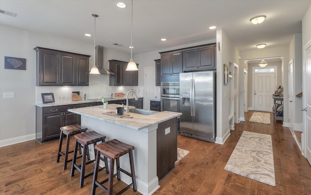 kitchen featuring dark hardwood / wood-style flooring, a kitchen island with sink, stainless steel appliances, wall chimney exhaust hood, and decorative light fixtures