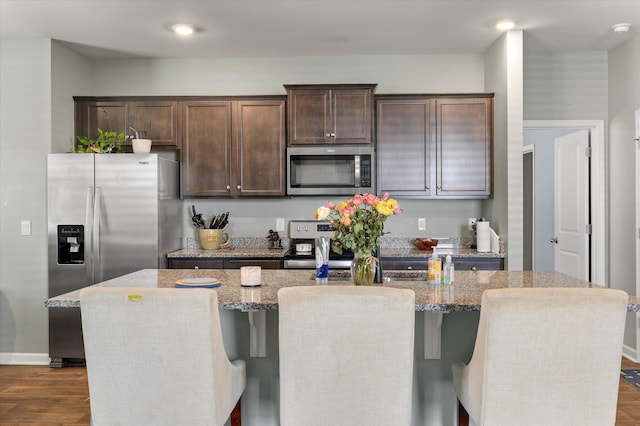 kitchen with dark brown cabinetry, dark wood finished floors, appliances with stainless steel finishes, a kitchen breakfast bar, and a kitchen island with sink