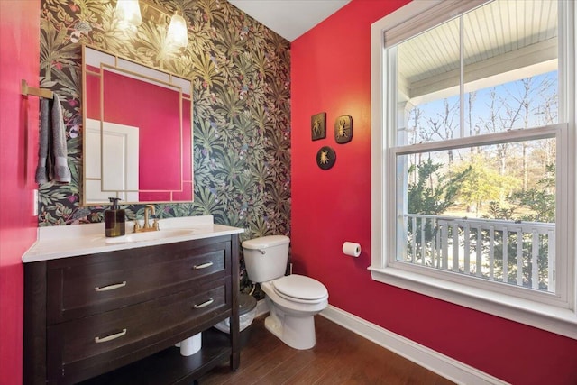 half bathroom featuring toilet, wood finished floors, wallpapered walls, baseboards, and vanity