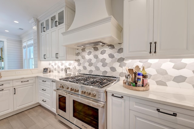 kitchen with premium range hood, range with two ovens, white cabinetry, light stone countertops, and decorative backsplash