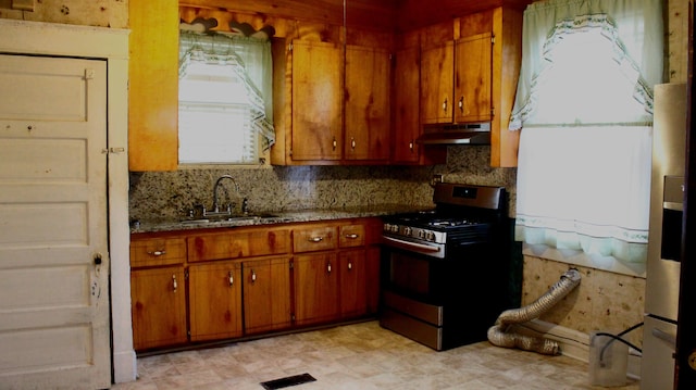 kitchen with brown cabinets, tasteful backsplash, a sink, under cabinet range hood, and stainless steel gas range oven