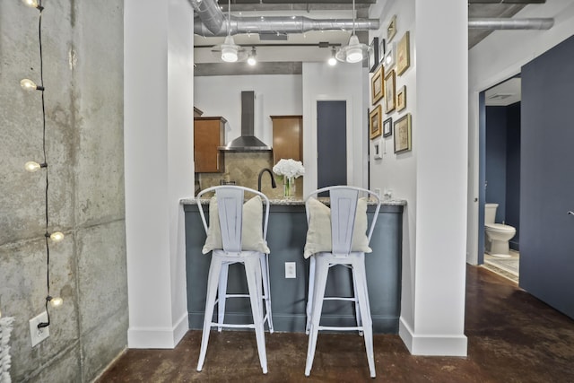 interior space with brown cabinets, backsplash, wall chimney range hood, concrete floors, and a kitchen bar