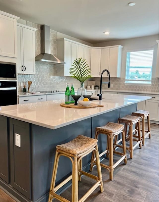 kitchen featuring wood-type flooring, stainless steel appliances, an island with sink, white cabinets, and wall chimney exhaust hood