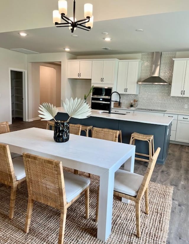 kitchen featuring wall chimney range hood, pendant lighting, an island with sink, white cabinets, and dark wood-type flooring