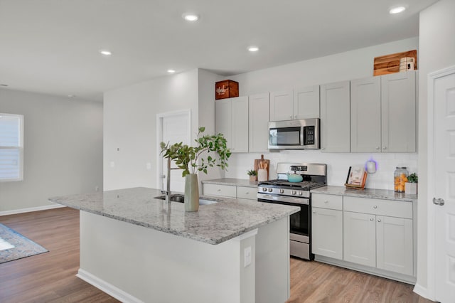 kitchen with light hardwood / wood-style floors, stainless steel appliances, light stone countertops, and an island with sink