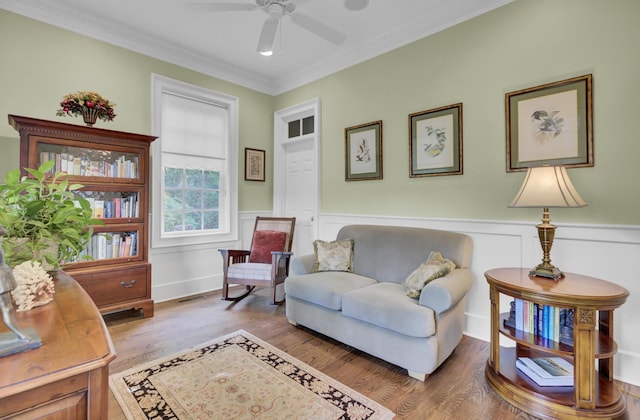 sitting room with wood finished floors, ceiling fan, ornamental molding, and a wainscoted wall