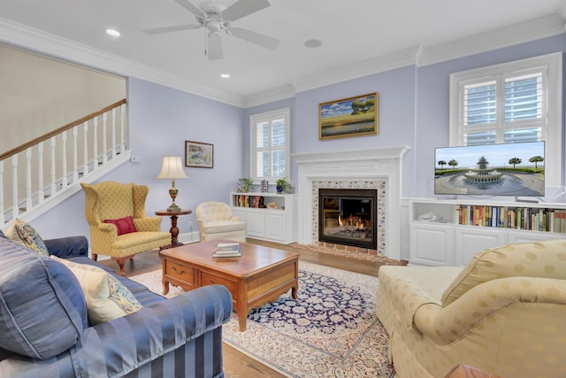 living room featuring wood finished floors, ornamental molding, ceiling fan, stairs, and a glass covered fireplace