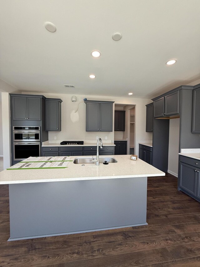 kitchen featuring a sink, dark wood-style flooring, gray cabinets, stainless steel double oven, and a kitchen island with sink