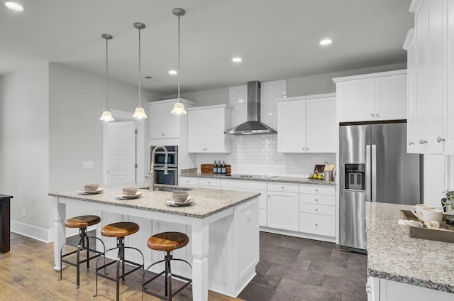 kitchen featuring a center island with sink, appliances with stainless steel finishes, white cabinetry, wall chimney range hood, and hanging light fixtures