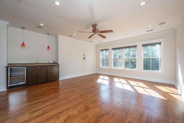 unfurnished living room featuring beverage cooler, ornamental molding, wood finished floors, bar area, and ceiling fan